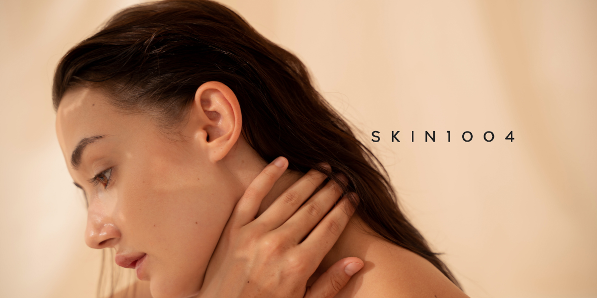 A close-up lifestyle shot of a woman with wet, dark hair touching her neck, set against a warm beige background with the text 'SKIN1004' in the upper right.